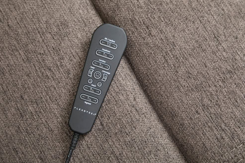 A black remote control with many buttons and white labels rests on the textured brown cushion of a Zecliner Model 3+ Umber Power Lift Recliner.