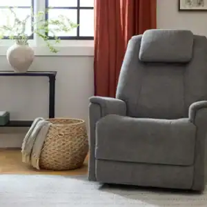 A Zecliner Model 3+ Umber Power Sleep Chair sits on a light rug in a cozy living room, next to a woven basket with a blanket, a console table with greenery, and a window with rust-colored curtains.