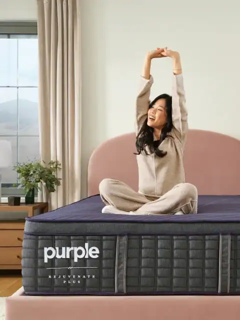 A smiling woman in pajamas sits cross-legged on a bed, stretching her arms up. The bed features a dark blue PurplePlus Mattress (Copy), with a nightstand and window in the background.