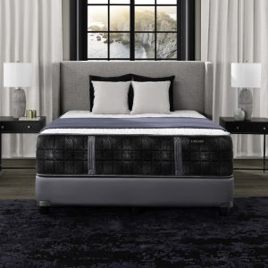 A neatly made bed with Summit Plush black and gray bedding sits between two black nightstands with white lamps, in front of large windows with a coastal view and light curtains framing the scene.