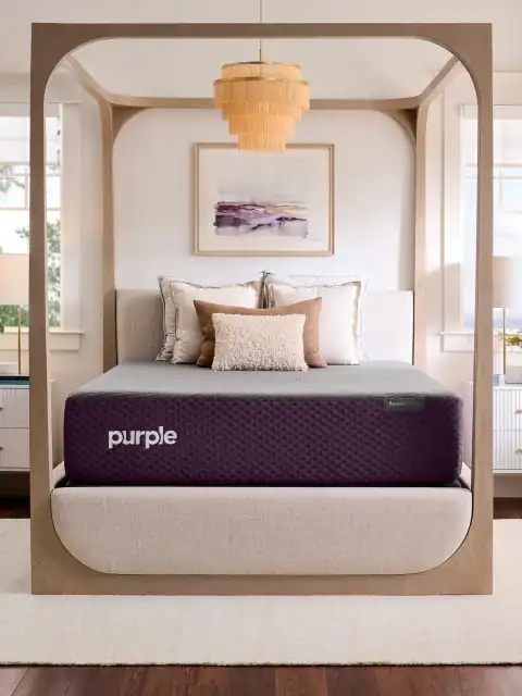 A bright, stylish bedroom features a modern four-poster bed with a Purple RestorePlus Hybrid Mattress (Copy), topped with decorative pillows. A chandelier hangs overhead, and a framed abstract painting decorates the wall behind the bed.