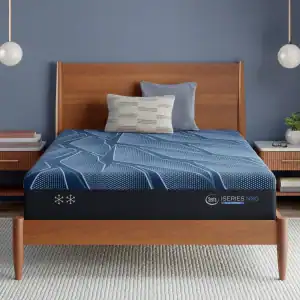 A modern bedroom with a wooden bed frame and nightstands, featuring a blue Serta iSeries NXG 3500 Hybrid — Firm mattress with geometric patterns, a gray pillow, and a patterned cushion against a blue wall.