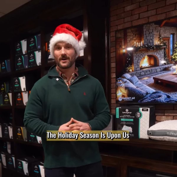 A man wearing a Santa hat and green sweater stands indoors by shelves of boxed products&mdash;perfect for last minute Christmas gifts. Behind him, a TV displays a cozy fireplace scene. On-screen text reads, "The Holiday Season Is Upon Us.