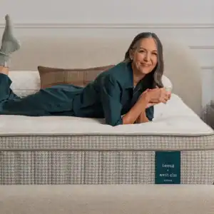 A woman in teal pajamas and gray socks smiles on a neatly made bed with a Leesa Reserve Hybrid Mattress in a cozy bedroom with a wooden nightstand and a plant in the background.