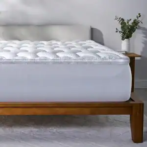 A wooden bed frame features a thick React Pro Performance Mattress Topper, complemented by a light upholstered headboard. Nearby, a small wooden side table holds a white vase with green leaves.