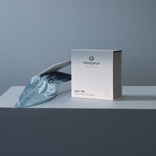 A pale blue Hyper Silk Performance Pillowcase partially slides out of a white box labeled "bedgear," resting on a minimalist blue-gray surface against a matching wall.
