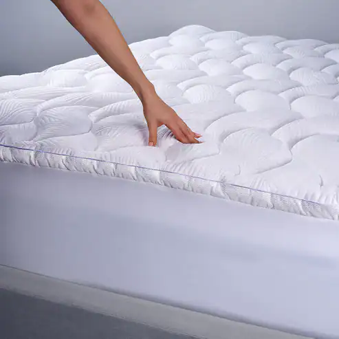 A hand presses on the React Pro Performance Mattress Topper, which features a white, quilted wave design and rests atop a neatly made bed with a white fitted sheet.
