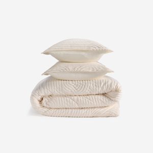 Two cream-colored pillows from the Performance Duvet & Sham Set - Racetrack are stacked on a matching quilt, all with a subtle, textured racetrack-inspired wavy pattern, set against a plain white background.