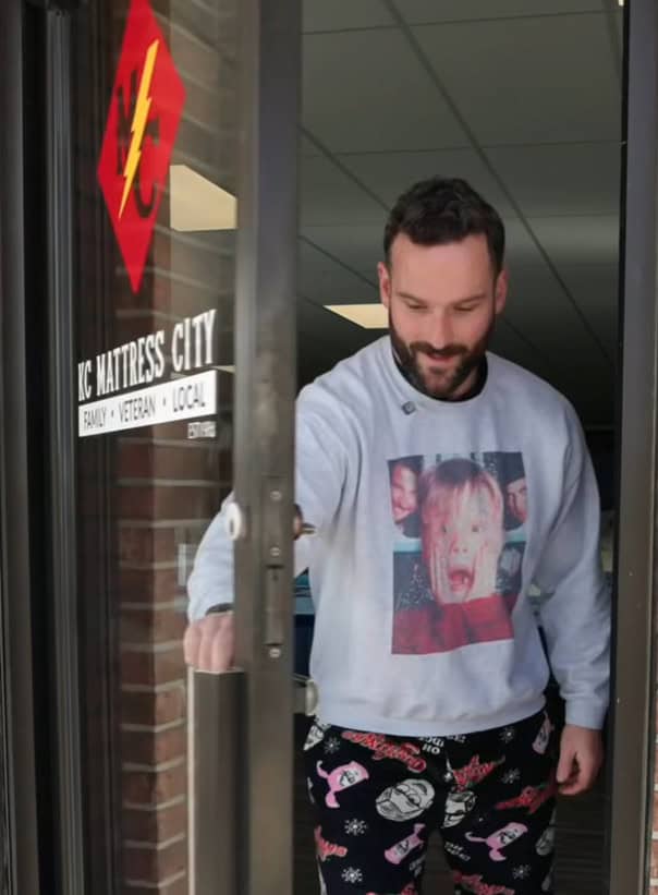 A man wearing a "Home Alone" sweatshirt and festive pajama pants opens the glass door of KC Mattress City, a shop with a sign reading "Family &bull; Veteran &bull; Local," ready to chat about Christmas plans.