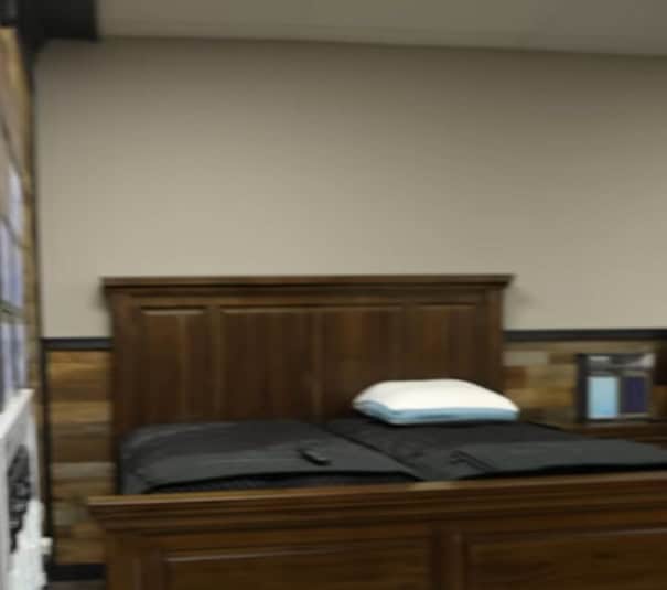 A wooden bed with a high headboard, two black mattress pads from KC Mattress City, and a single white pillow sit in a room with tan walls and wood panel accents. A small framed picture hangs on the right wall above a nightstand.