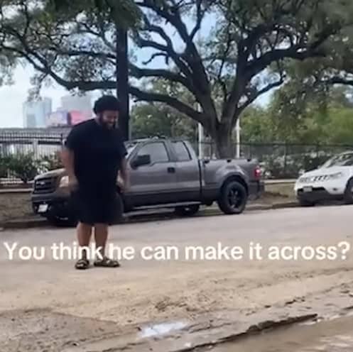 A man stands on a street, preparing to cross a wide puddle. Parked cars and trees are in the background. Text on the image reads, "You think he can make it across?.