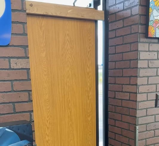 A wooden sliding door is partially open inside a building with brick-patterned walls. Colorful artwork and a window showing the outdoors add charm to the scene, perfect for showcasing special Black Friday d&eacute;cor deals.