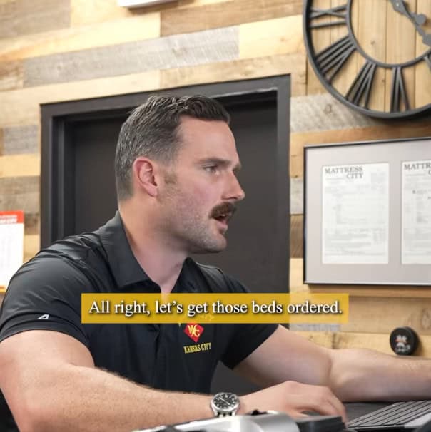 A man with a mustache and short hair sits at a rustic office desk, speaking. Text reads, "All right, let's get those 6 or 7 beds ordered." A large wall clock hangs on the wall behind him.