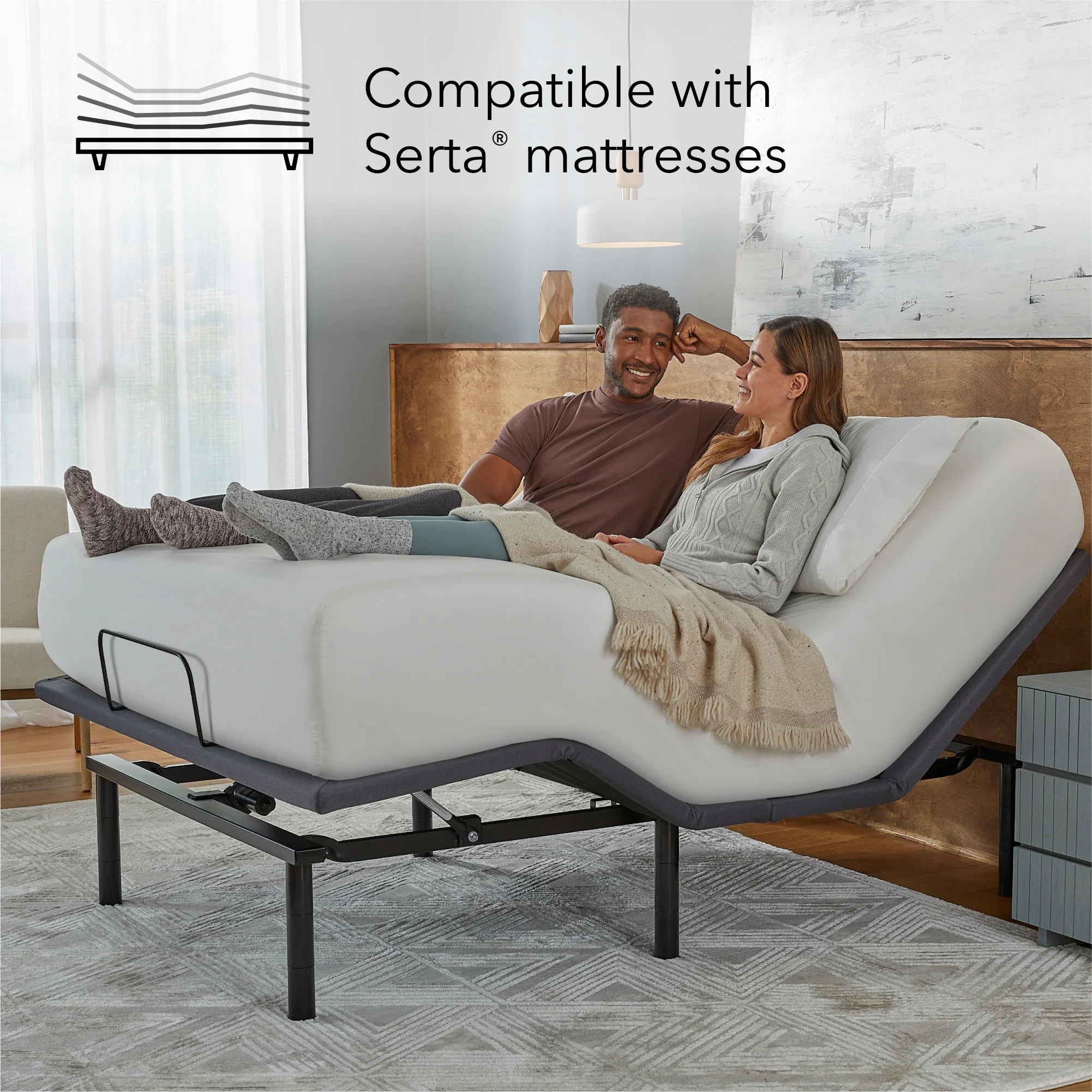 A smiling couple relaxes on a Baselogic™ Silver Adjustable Base with raised head and foot sections in a cozy bedroom. Text reads, “Compatible with Serta® mattresses.”.