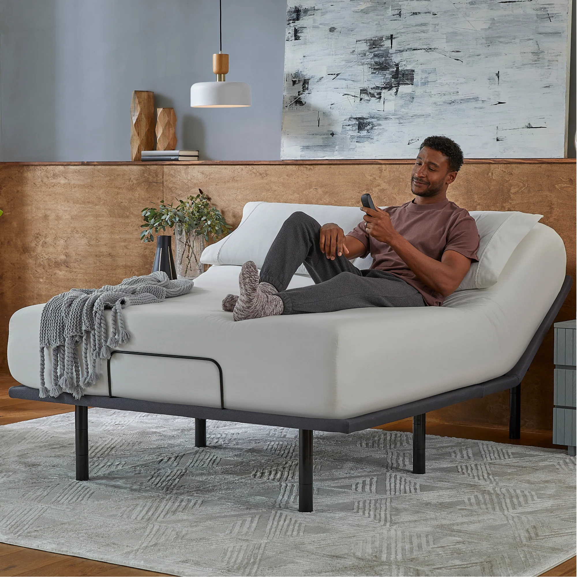 A man in comfortable clothes relaxes on the Baselogic™ Silver Adjustable Base with raised head and knees, using a remote. The modern room features a rug, plants, and abstract wall art, highlighting Baselogic™ comfort.