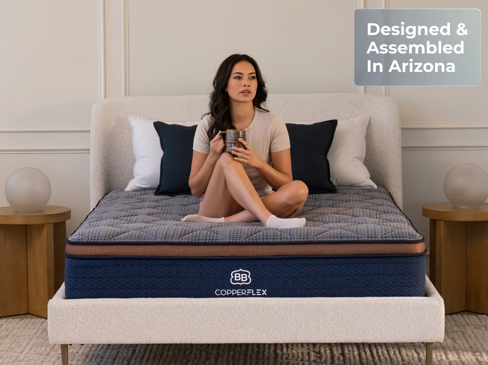 A woman sits on a bed holding a mug, with navy and white pillows behind her. The CopperFlex Hybrid 12" mattress is labeled "CopperFlex." A sign in the corner reads "Designed & Assembled in Arizona.