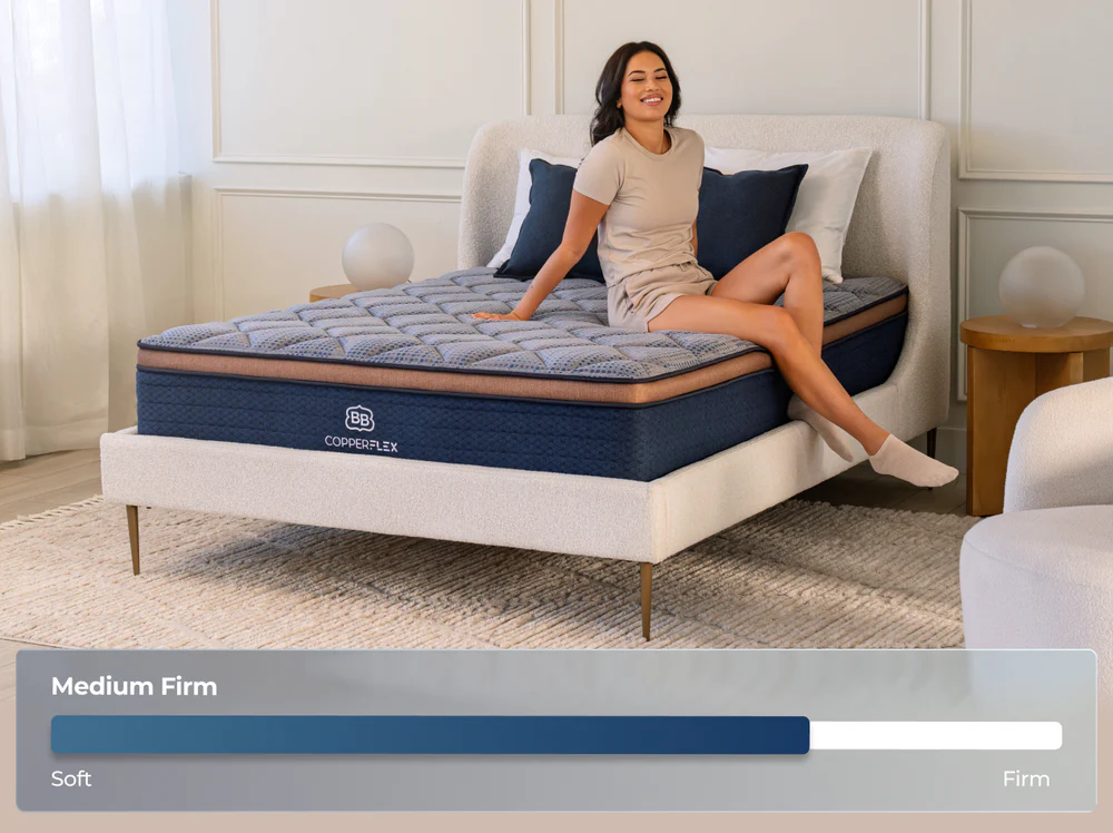 A woman smiles on a modern upholstered bed with the CopperFlex Hybrid 12" mattress in blue and gray. A graphic shows it's medium firm. The room has light walls and elegant minimalist decor.