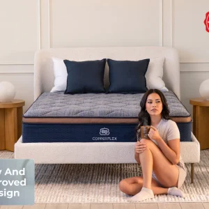 A woman sits on the floor, leaning against a bed with a CopperFlex Hybrid 12" mattress, holding a mug. The room has neutral decor, wooden side tables with lamps, and a "New And Improved Design" label in the corner.