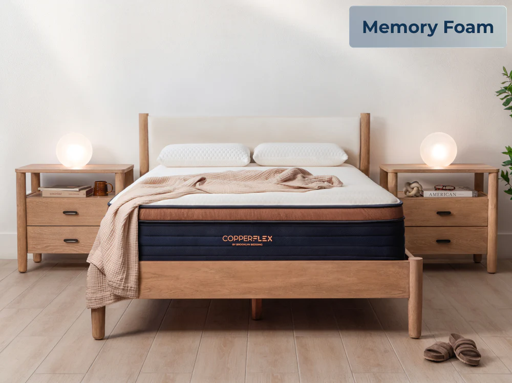 A neatly made bed featuring the CopperFlex Pro Hybrid 14" mattress with beige bedding sits between two wooden nightstands, each topped with a glowing round lamp, in a bright minimalist bedroom. A "Memory Foam" label appears in the corner.