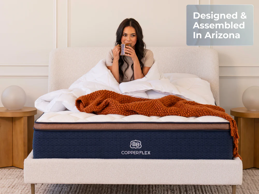 A woman relaxes in bed, sipping from a mug, wrapped in a white comforter and rust-colored blanket on the CopperFlex Pro Hybrid 14" mattress. A sign beside her reads, "Designed & Assembled In Arizona.