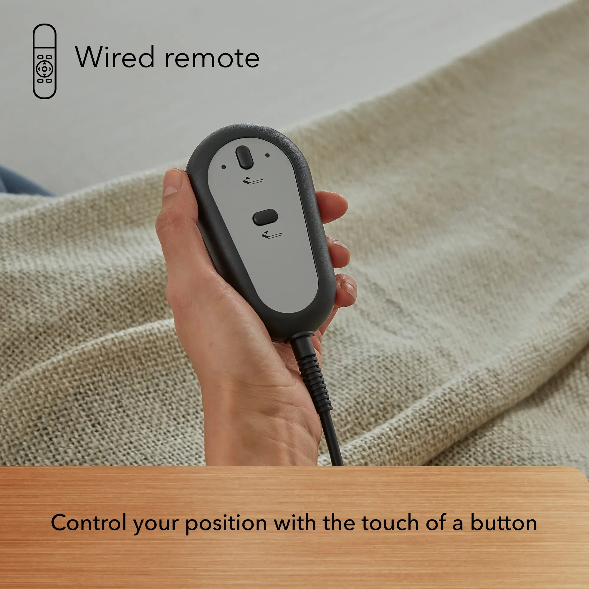 A hand holds a wired remote with two arrow buttons over a beige textured blanket. Text above: "Wired remote." Below: "Easily adjust your Baselogic™ Bronze Adjustable Base position with the touch of a button.