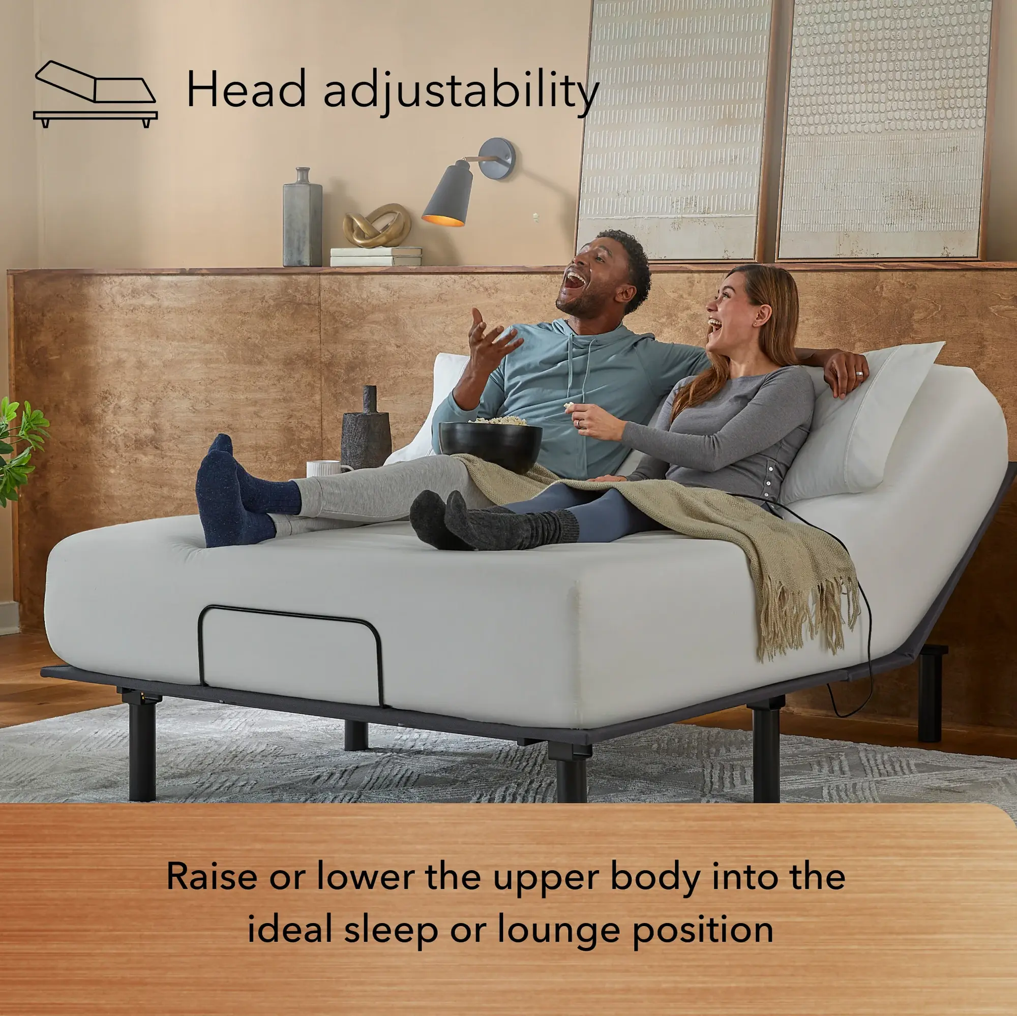 A smiling couple relaxes on a Baselogic™ Bronze Adjustable Base with the head section elevated in a modern bedroom with warm brown tones. Text: "Head adjustability—raise or lower your upper body for ideal sleep or lounging.