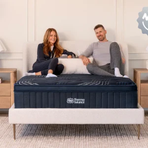 A smiling woman and man sit on a ThermoBalance Soft mattress in a bright, modern bedroom with a light headboard, two nightstands with lamps, and a "NEW" label in the top right corner.