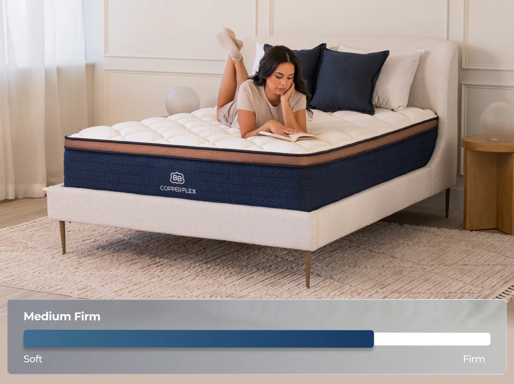 A woman reads on her stomach atop a neatly made CopperFlex Pro Hybrid 14" mattress, displayed on a comfort scale from soft to firm in a softly lit, modern room with neutral, minimalist decor.